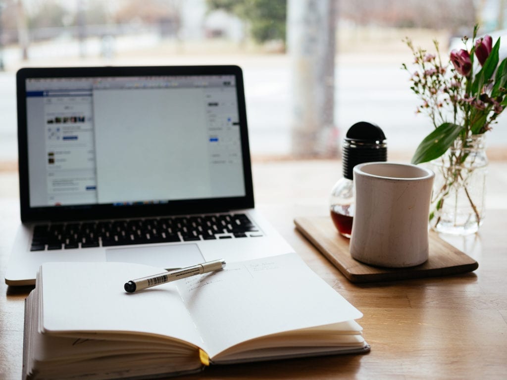 laptop, open book with pen, and coffee on sunny desk