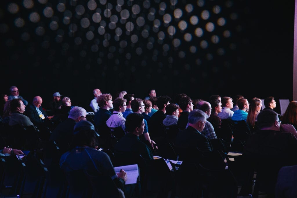 people in darkly lit room at a conference taking notes listening to a speaker